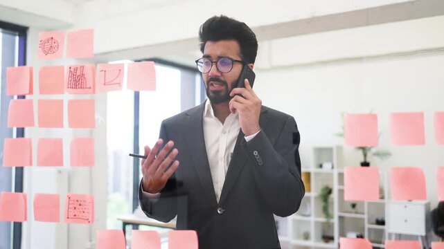 An Indian businessman wearing glasses talks on his phone while looking at sticky notes on a glass wall