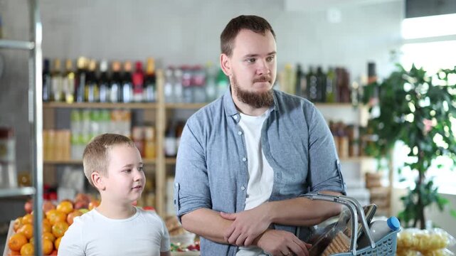 Visiting store, father and son is looking at something on shelf of showcase. Family holds heavy basket of groceries and reaches for shelf, trying to see products in refrigerator window.