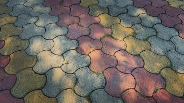A slow tracking view over patterned interlocking tiles in red, yellow, and gray, softly lit by shifting tree shadows that create gentle contrasts on the ground.