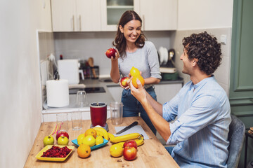 Happy smiling couple share a moment together while preparing homemade fruit drinks, with a countertop full of oranges, apples, and bananas.