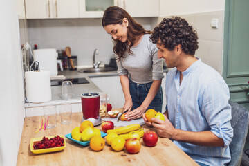 Smiling and relaxed, the two share a moment together while preparing homemade fruit drinks, with a countertop full of oranges, apples, and bananas.