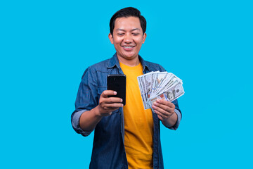 Excited Asian Man Holding Cash and Smartphone Celebrating Financial Success Against Blue Background