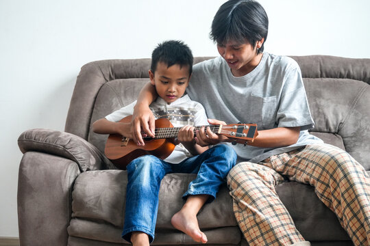 Portrait Of Two Asian Brothers Playing Ukulele Together While Sitting On Sofa - Powered by Adobe