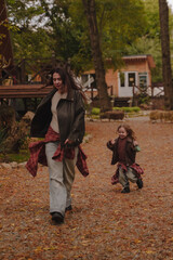 Obraz premium Mother and daughter run around the autumn yard near cozy A-shaped houses. The girls appear to be dressed in leather jackets, blue jeans and plaid shirts. Autumn atmosphere. Vertical photo.