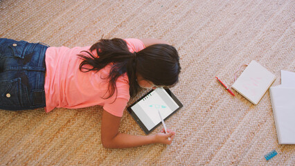 Overhead Shot Of Young Girl Lying On Floor in Lounge At Home Drawing With Stylus On Digital Tablet