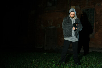 Criminal man in hood with gun near brick building outdoors, space for text