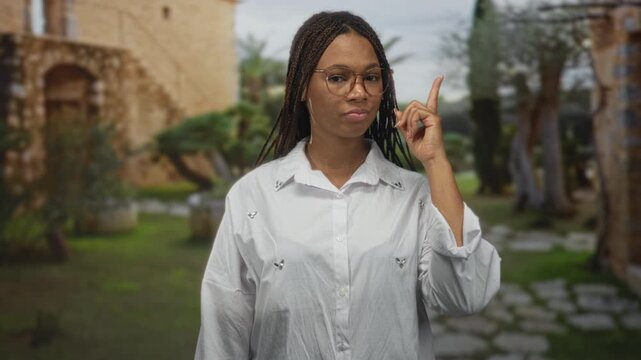 Woman pointing finger at camera in building courtyard with old stone facade; confidence assertiveness invitation.