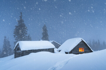 Fototapeta premium Cozy house with warm light during winter snowfall in a snowy forest. Cold mountain night landscape with snow covered trees