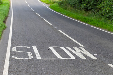 White road marking reading SLOW painted on a rural asphalt road with grass verges. Concept of traffic safety, caution and speed awareness