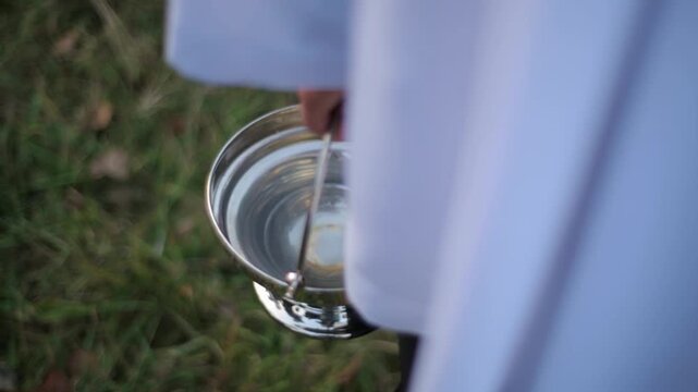Close-up of a metal container (bucket) holding transparent holy water, held by a young ministrant or altar server dressed in white liturgical vestments outdoors. Symbolizes baptism, blessing, purifica