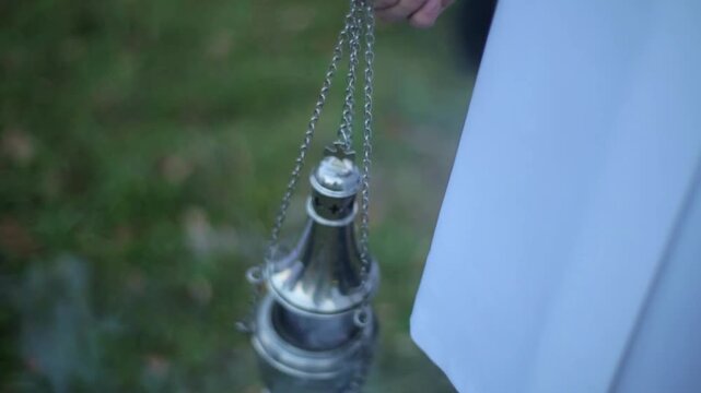 Close-up of a silver metal thurible or censer hanging from chains, actively swinging and emitting thick incense smoke during a religious or commemorative service outdoors.