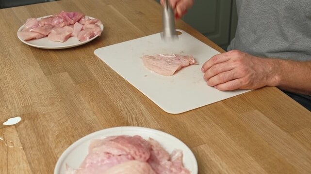 Person using meat tenderizer on raw chicken. Chef preparing pork loin slice with meat mallet on cutting board. Man tenderizing fillet. Cooking cutlets pounding poultry breast close-up. Culinary dish.