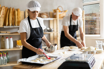 Two bakers prepare dough for baking - they put pieces of raw dough on baking sheet and pour oil over them