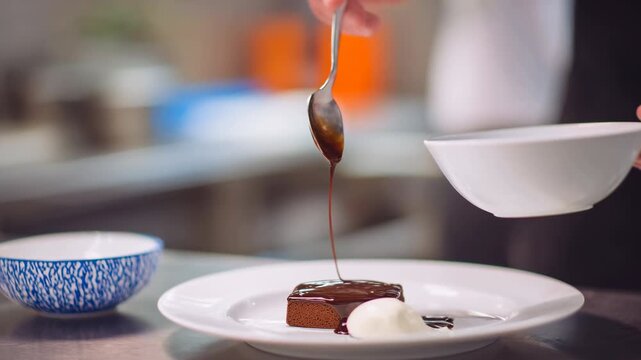 Rich chocolate sauce poured over brownie with cream plated dessert professional kitchen spoon pouring glossy chocolate cake sauce over slice white