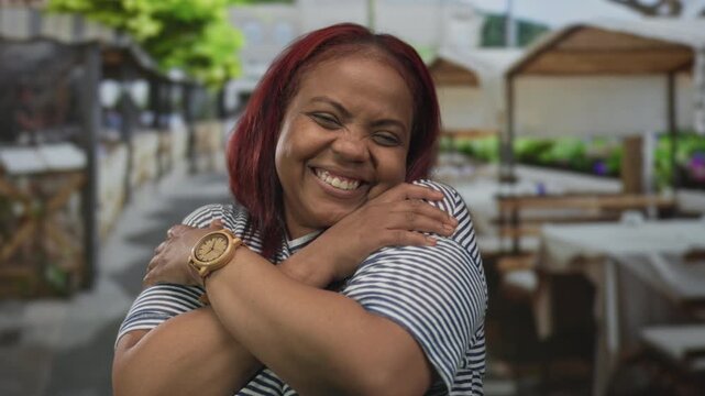 Woman smiling warmly hugging herself on a sunny outdoor restaurant terrace with wooden tables visible; joy self love comfort.