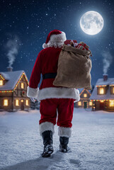 Santa, seen from the back, carries a large sack of presents over his shoulder while walking away through a snowy village illuminated by a full moon and twinkling lights.