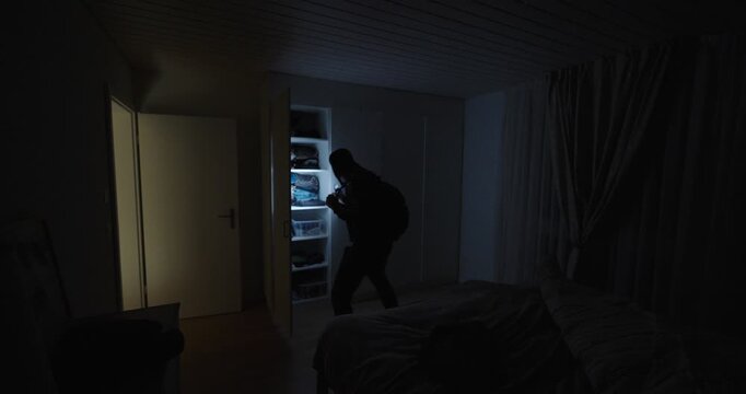 A masked intruder moves through a dark bedroom using a flashlight to search shelves and drawers, symbolizing burglary, home invasion, theft, and nighttime danger.