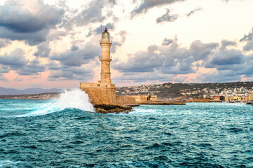 Old Venetian Lighthouse in Chania Crete hit by powerful turquoise wave under dramatic pastel sky. Scenic harbor view with dynamic sea motion, stone pier, warm evening light, stormy coastal atmosphere.