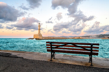 Wooden bench overlooking Venetian Lighthouse in Chania Crete Greece during calm evening with pastel clouds, turquoise sea, peaceful harbor scenery, tranquil coastal mood, inviting Mediterranean atmosp