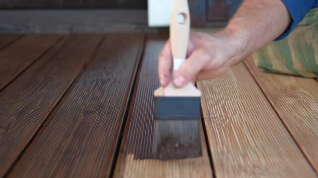 Brown oil for protection of wood by staining. Painting a larch for protecting and pests. Woodworker applying finish to terrace deck board. Repairman varnishing with brush wooden plank floor surface.