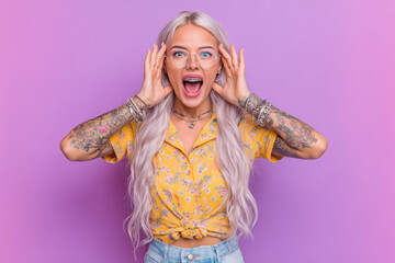 Excited woman with long blonde hair and tattoos showing surprise against a purple background, wearing glasses and a yellow shirt, expressing joy