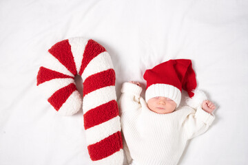 A cute baby sleeps in a Santa hat against a white background with red candy canes. The newborn is celebrating his first Christmas.