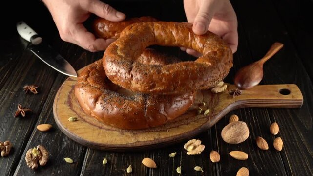 Hands are placing freshly baked bread on a wooden board. Surrounding the bread are various nuts and a knife, indicating preparation for a meal in a kitchen.
