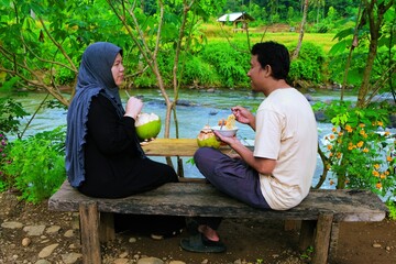 A couple enjoying a relaxed conversation by the riverside, surrounded by nature and peaceful rural scenery.