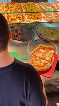 Customer Choosing Rectangular Pizza From Heated Display Counter, Server Handing Tray, Bright Display Lighting And Warm Slices Visible, Takeaway Ordering Scene. Vertical shot taken on mobile device