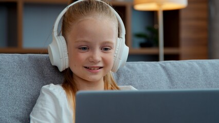 Caucasian smiling little child girl in headphones looking laptop at home happy kid daughter e-learning studying online watch lesson listen audio music playing computer game watching movie cartoons