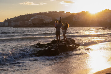 A father raises his arm in joy while standing with his family on a rock amidst crashing waves at sunset in Almuñécar, celebrating a holiday evening on the Costa Granadina beach in Andalusia, Spain.