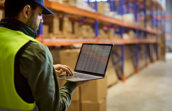 Warehouse inventory worker uses laptop for stock. He checks goods on shelves and updates storage records to support logistics, audit and management. Clear concept of inventory management.