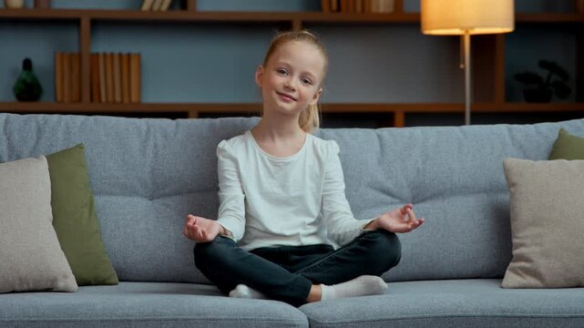 Healthy little Caucasian girl meditate smiling looking at camera at home kid daughter practicing meditation sitting in lotus pose on couch meditating preteen child stress relief breathing exercise