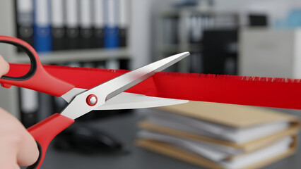 Hand holding red scissors cutting a red ribbon, symbolizing the opening of an event or business in an office setting with stacks of files