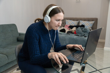Focused young woman wearing headphones works from home, typing on her laptop and using a mouse,...