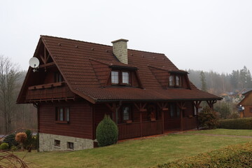 Obraz premium Traditional Wooden Cottage with Red Tiled Roof and Stone Chimney, Rustic Mountain House Exterior with Balcony and Porch, Rural Architecture in Foggy Weather, Classic Timber Home Facade