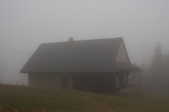 A traditional wooden mountain cottage (chalupa) barely visible in thick, heavy fog. A mysterious and isolated rural house scene, conveying solitude and quiet.