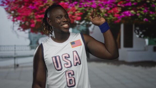 Young man in usa basketball jersey smiling and holding up peace sign with bare arm and blue wristband beside building and flowering trees; joy patriotism.