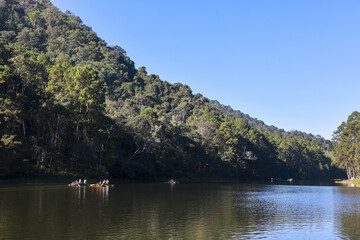 Serene landscape of calm lake with lush green forest mountain under clear blue sky. People on bamboo rafts enjoy peaceful nature and tranquil water reflection