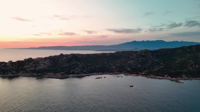Cinematic aerial pan over rugged Capu di Fenu coastline near Bonifacio, Corsica, with distant mountains silhouetted against a dramatic golden hour sunset sky.