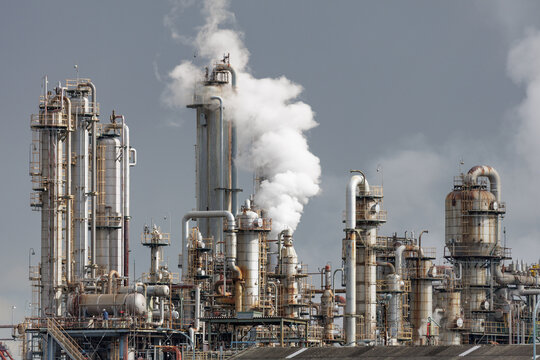 Refinery emits white steam plumes that rise over a distillation tower matrix in sprawling vintage industrial complex set on overcast sky backdrop.