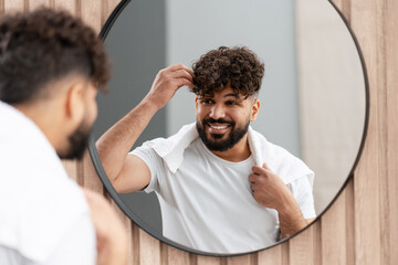 Young man smiling, adjusting curly hair in bathroom mirror, enjoying morning routine