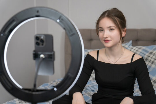 Young smile influencer sitting on bed, recording video content with smartphone and ring light, creating engaging material for her online audience - Powered by Adobe