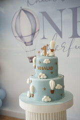 A dreamy two-tier blue birthday cake decorated with hot-air balloons, fluffy clouds, and golden stars. Designed for Khalid&rsquo;s first birthday, it creates a soft, whimsical, sky-themed celebration moment