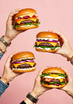 Four hands holding cheeseburgers with beef, bacon, egg, lettuce, tomato, and sauce against a light pink background.