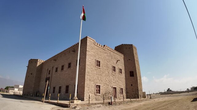 Mirbat fort. A historical stone structure with battlements and corner towers. Stands under a clear blue sky. Proudly flying the Omani flag. Representing national heritage and strength in Dhofar region