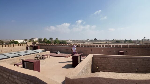 Woman walking along Mirbat Fort s ancient stone walls in Dhofar, Oman, overlooking the Arabian Sea and coastal city under a clear blue sky during sunny travel day in Mirbat town