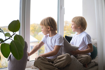 children, blonde twin boys, look out the window, sitting on the window sill