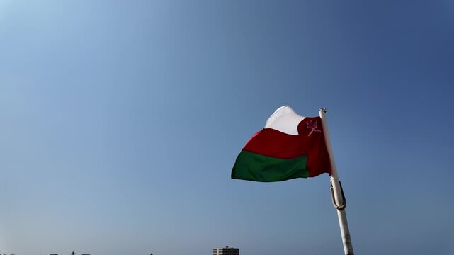 Oman flag flying in slow motion from a flagpole over Mirbat, Dhofar, waving against a clear blue sky above the Arabian Sea, symbolizing national pride, heritage of Oman in Mirbat town