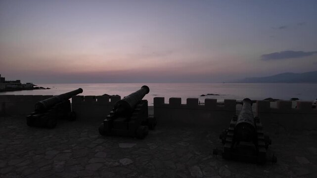 Ancient cannons positioned on the historic Mirbat fort wall at sunset, overlooking sandy beach and turquoise waters of the Arabian Sea in Dhofar, Southern Oman, showing coastal defense in Mirbat